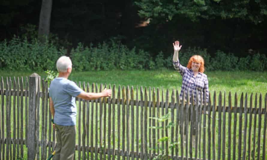 man with white hair back to screen, with his hands on his backyard fence talking to his neighbour, a lred hair smiling lady with a raised right arm waving at him for the secret to happiness